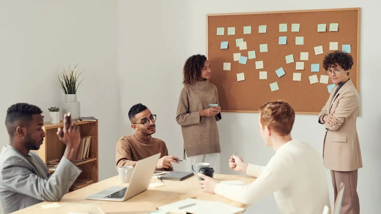 A diverse group of people collaborates in an office, with a bulletin board displaying various notices in the background.
