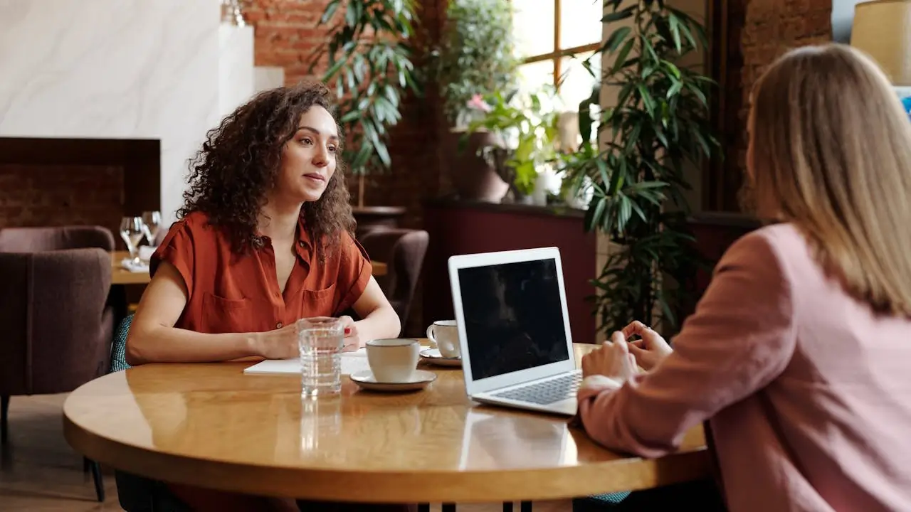 Two women seated at a table, collaborating on a laptop, engaged in discussion and sharing ideas